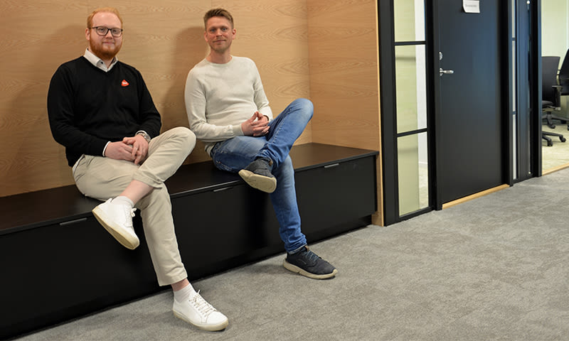 To menn sitter på svart benk mot lyst trepanel, grått gulv, dør med vindu i kontorlandskap