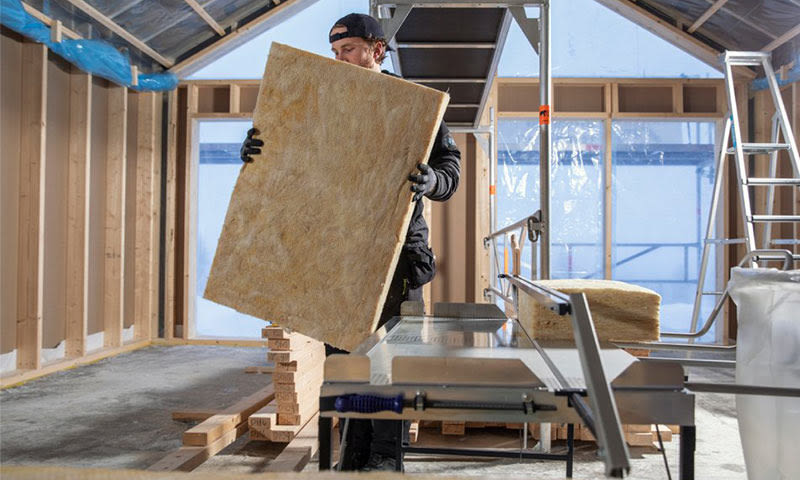 Arbeider i råbygg bærer gul isolasjonsplate foran sagbord og trestendere.