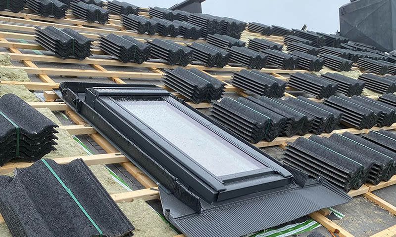 Skråtak under bygging med stabler av svarte takstein på lekter og nytt takvindu.