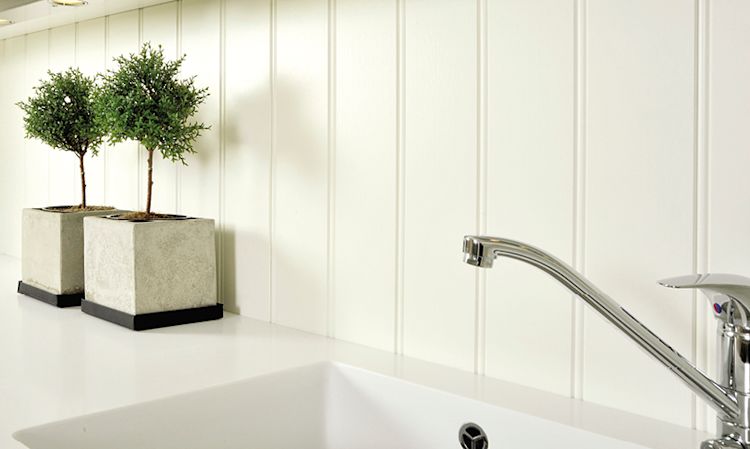 Hvit kjøkkenbenk med synlig vask og kromkran, hvit panelvegg og to små prydtrær i grå betongpotter.