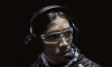 Person med mørk hettegenser og store svarte hodetelefoner mot svart bakgrunn.