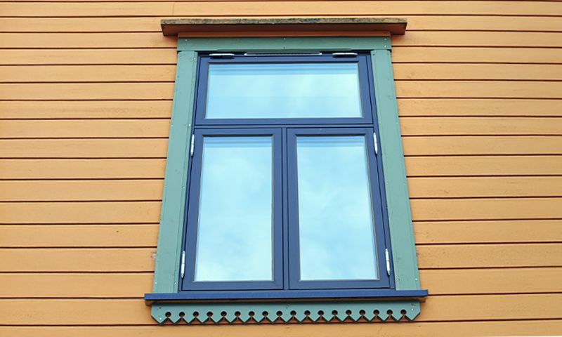 Vindu med blå ramme og dekorativ grønn list på gul trefasade, reflekterer blå himmel.