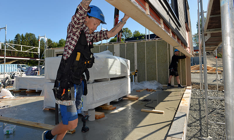 Bygningsarbeidere i blå hjelmer og verktøybelter løfter veggpanel på byggeplass i solskinn