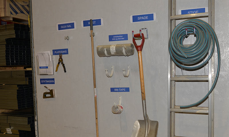 Verkstedsvegg med blå etiketter, platesaks, kost, spade, teip og stige med grønn slange