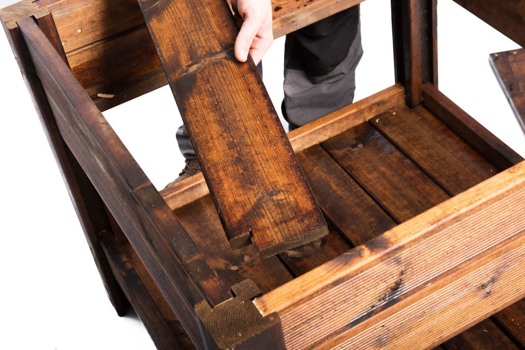 Hender setter en mørk brun treplanke i rustikk trekasse mot hvit bakgrunn.