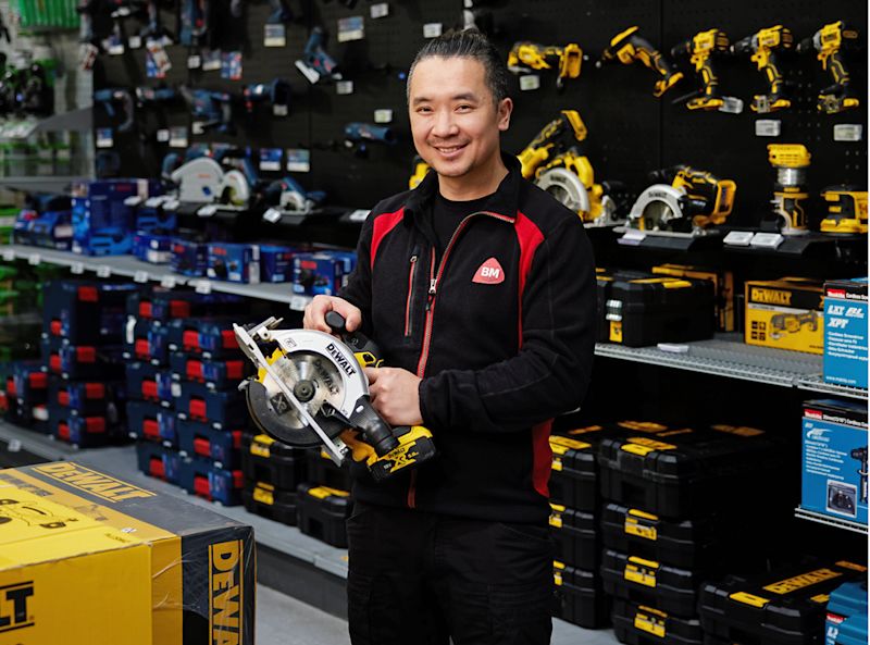 Ansatt i svart-rød jakke holder gul-svart DeWalt sirkelsag foran verktøyhylle.