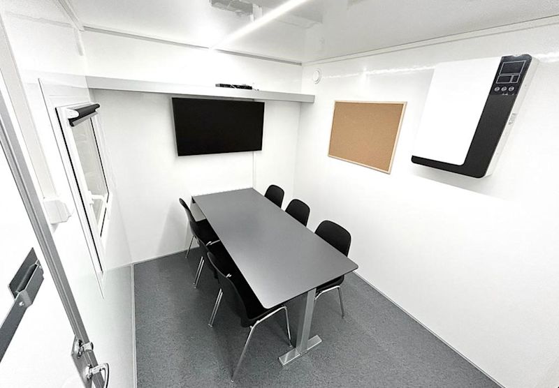 Lite hvitt møterom med grått bord, seks svarte stoler, vegg-TV, korktavle og luftrenser.