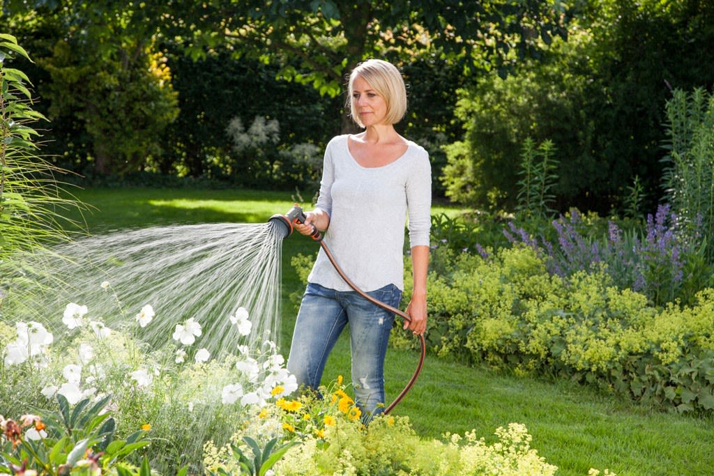Person med grå genser og blå jeans vanner hvite og gule blomster i solrik hage.