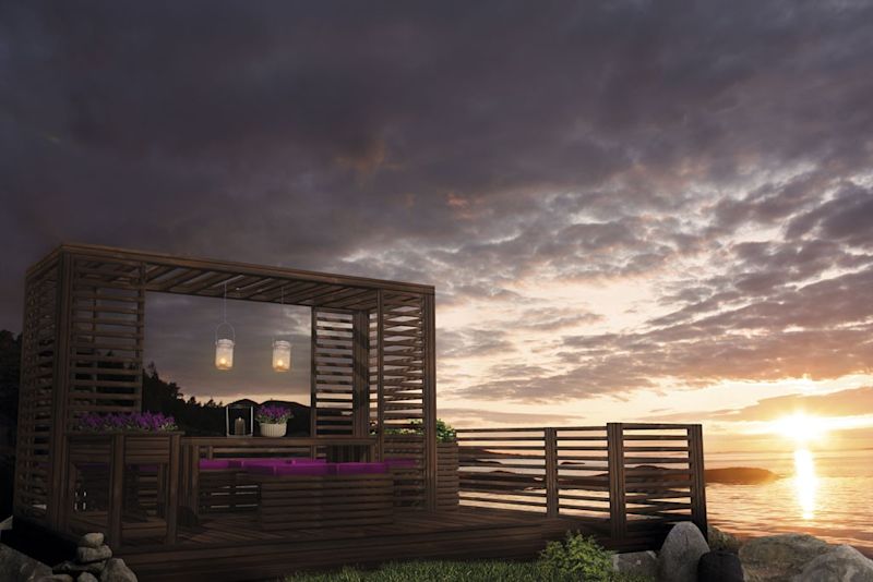 Trepergola med lanterner, lilla puter og blomster ved steinete strand i solnedgang