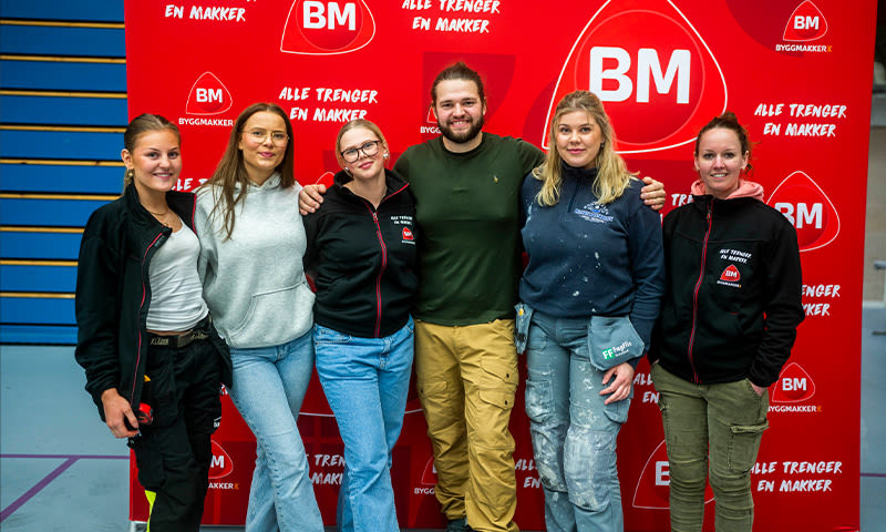 Seks personer i arbeidsklær foran rød BM-plakat med påskrift “Alle trenger en makker”