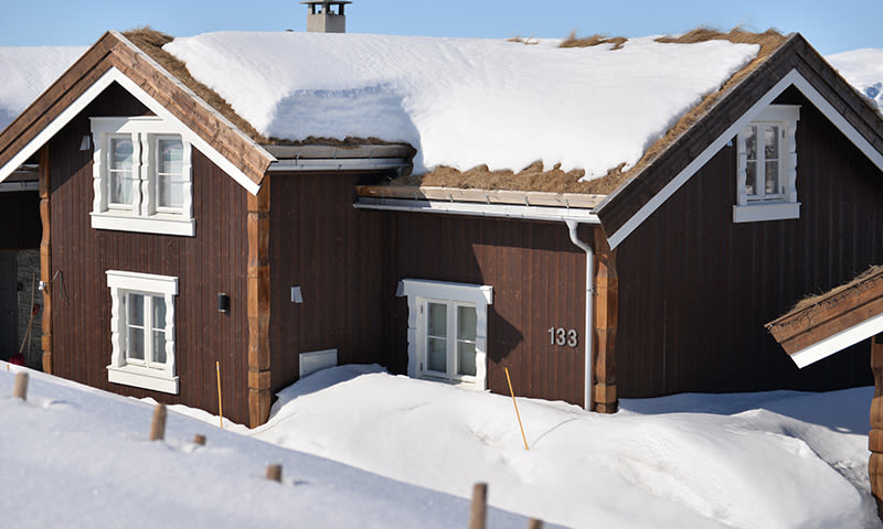 Brun tømmerhytte med hvite vinduskarmer under snøtykt tak og snødekt bakke, husnr.133
