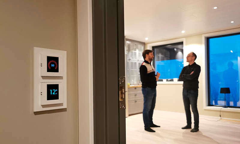 To digitale termostater (rød/blå) på gangvegg, to menn snakker i lyst parkettrom med vindu