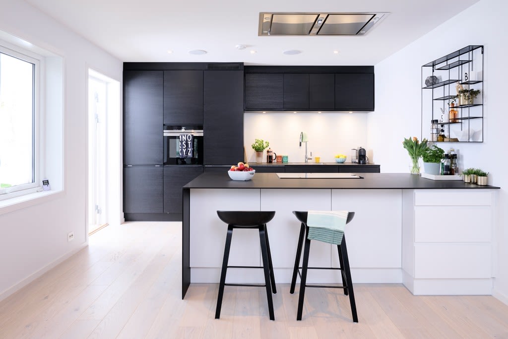 Lyst kjøkken med hvit øy, svart benkeplate, sorte overskap, to barstoler og vindu