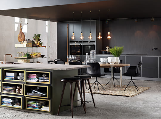 Moderne kjøkken i grått og grønt med lys steinbenk, hylle med bøker, svarte stoler og hengelamper.