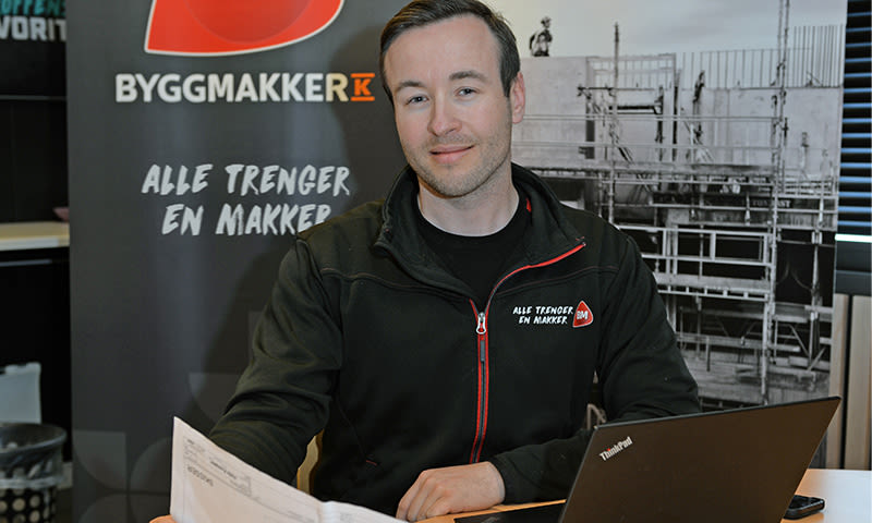 Mann i svart og rød Byggmakker-jakke ved ThinkPad-laptop foran sort banner og byggeplassbilde