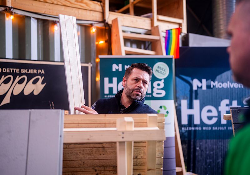 Mann holder treplanker ved en Moelven-stand med skilt og regnbueflagg i bakgrunnen.