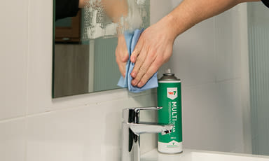 Hender tørker speil med blå klut ved hvit servant, grønn rengjøringsspray ved kromkran