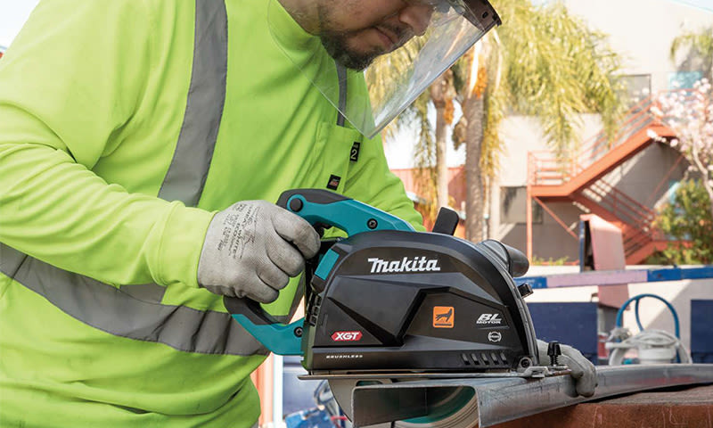 Bygningsarbeider i neongrønn refleksvest bruker svart-blå Makita XGT-metallsag på byggeplass