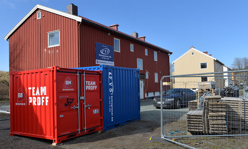 Byggeplass med rød trebygning bak rødt og blått container, gjerdestativ, paller og bil.
