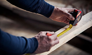 Hender som måler en treplanke med svart-rødt målebånd og gul stripe