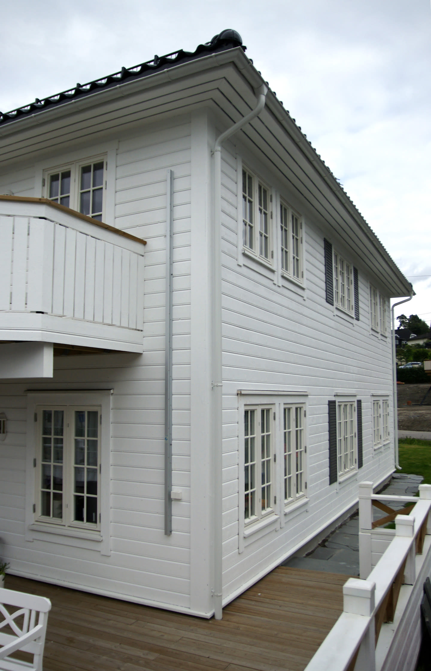 Hvitt toetasjes trehus med sprossevinduer, balkong, mørkt tak og treterrasse under overskyet himmel