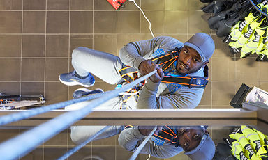 Arbeider i grå klær og blå hjelm klatrer i oransje sele og tau over grått flisgulv ved gule vester.