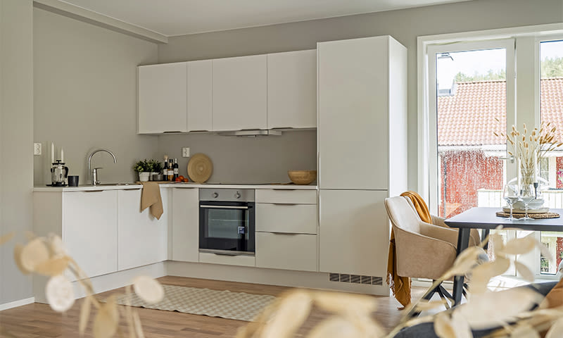 Lyst, moderne hvitt L-kjøkken mot grå vegg med stekeovn, gryter og spisebord ved vindu