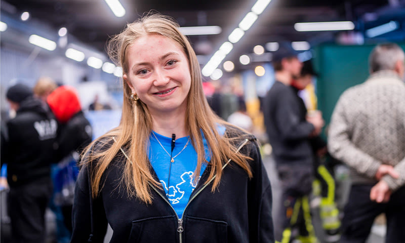 Ung kvinne i blå t-skjorte med Europa-kart og svart hettejakke i en lys messehall.