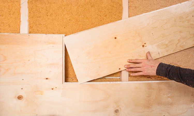 Hånd plasserer lyst kryssfinerpanel foran brunt isolasjonsmateriale på vegg.