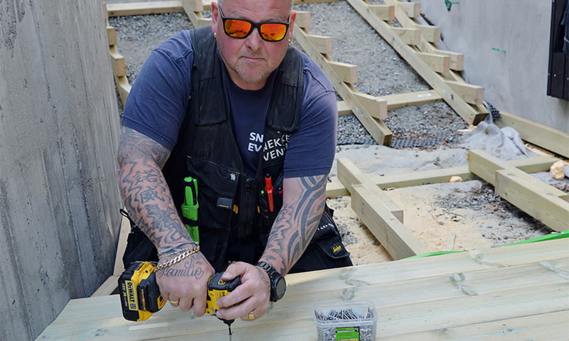 Tattooert håndverker skrur lyst tredekke med gul drill på byggeplass.