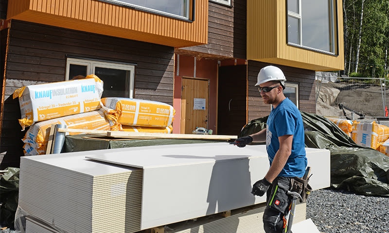 Byggearbeider i hjelm ved stabel hvite gipsplater foran trefasade med gule og brune paneler.