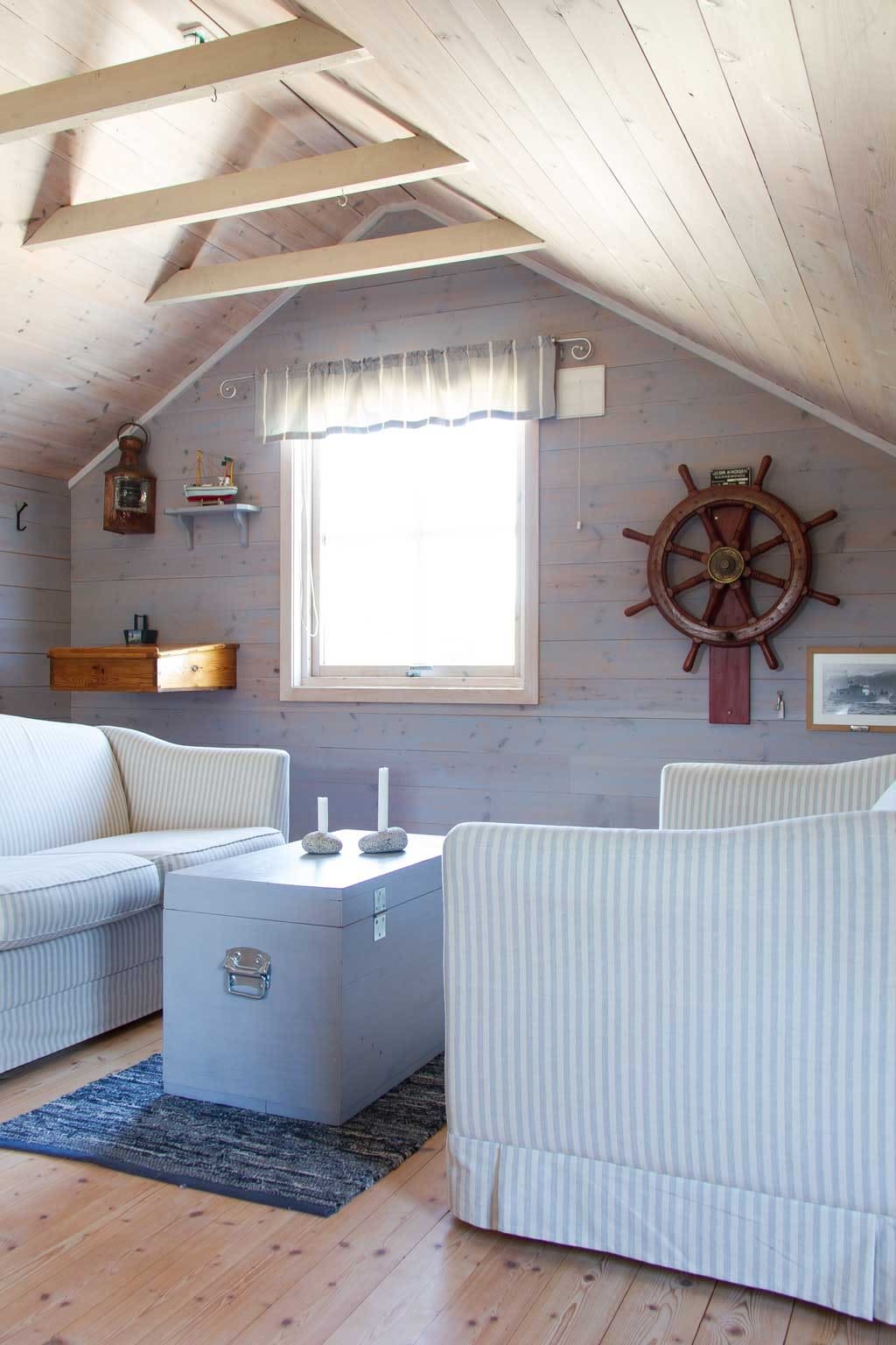 Lyst loftstue med lysegrå stripete sofaer, grå kistebord, stearinlys og skipshjul på trepanel.