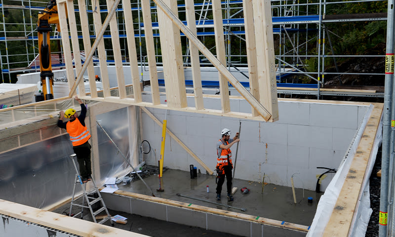 To arbeidere i oransje vester og hvite hjelmer løfter prefabrikert veggstamme på byggeplass