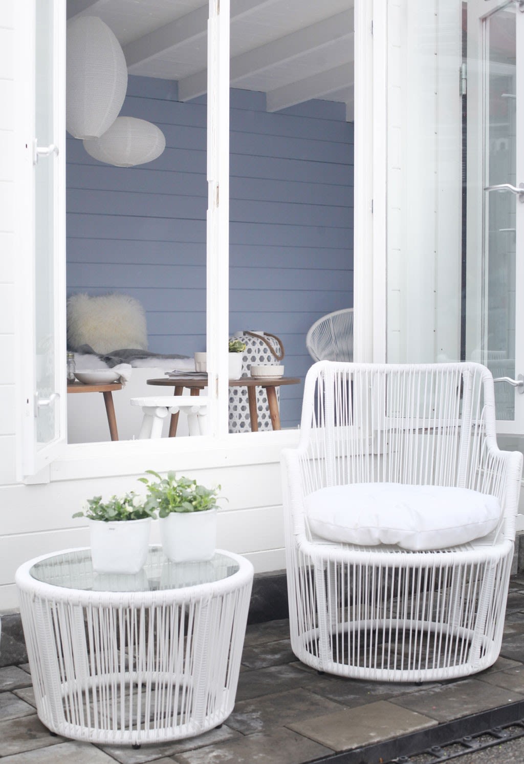 Hvitt flettet utebord og stol med pute foran åpent vindu mot blå vegg, to grønne potteplanter.