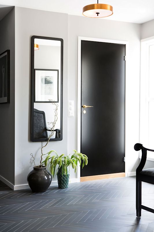 Lysegrå entre med svart dør, fiskebeinsgulv, høyt speil, grønne planter og gulllampe.