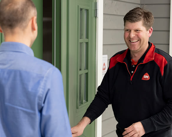 En mann i svart-rød BM-uniform står ved en åpen grønn dør og hilser en kunde i blå skjorte