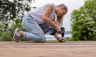 Person i hvit t-skjorte og blå jeans skrur med blå batteridrill på trebrygge, grønn hage bak