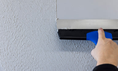 Hånd med blå sparkel fjerner ujevn gips fra grå, strukturert vegg under metalllist