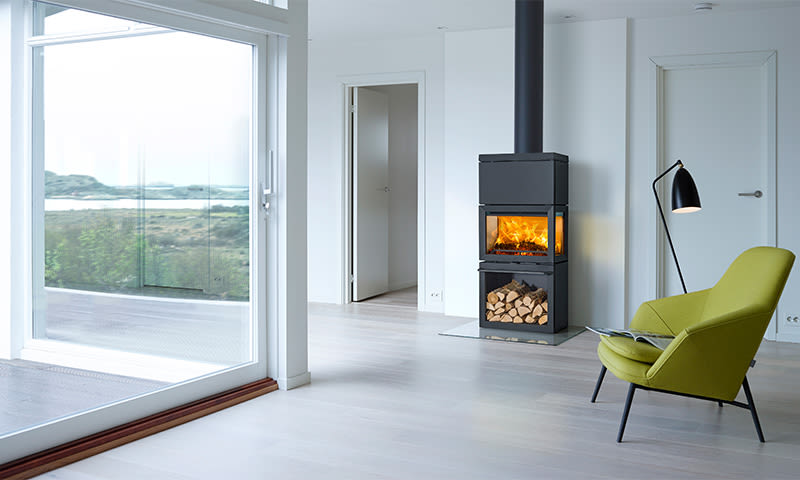 Lyst rom med svart ovn med ild, stablet ved, grønn lenestol, gulvlampe og vindu mot kystlandskap.