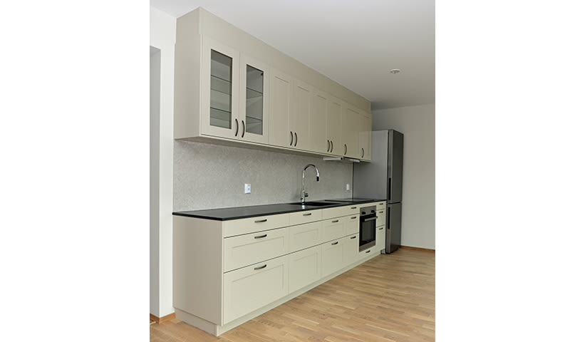 Lyst beige kjøkken med sorte benkeplater, stålvask, innebygd ovn, kjøleskap og eikeparkett.