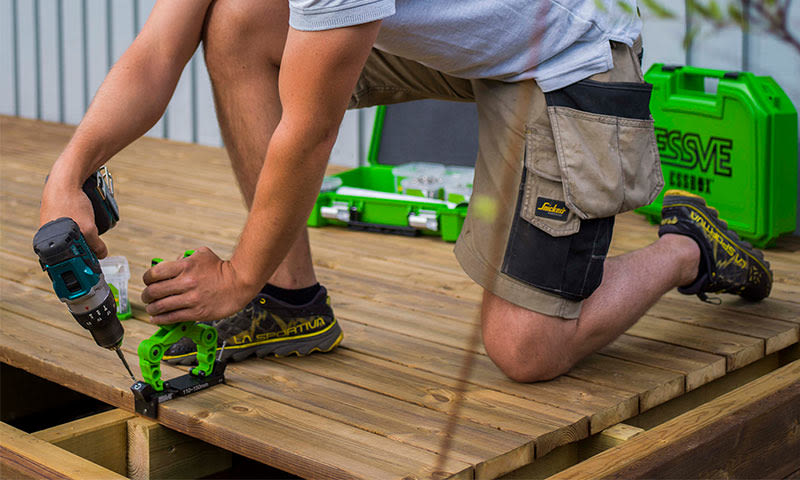 Mann knelende på brun terrasse, fester bord med grønn klemme og blå drill, grønn verktøykasse bak.
