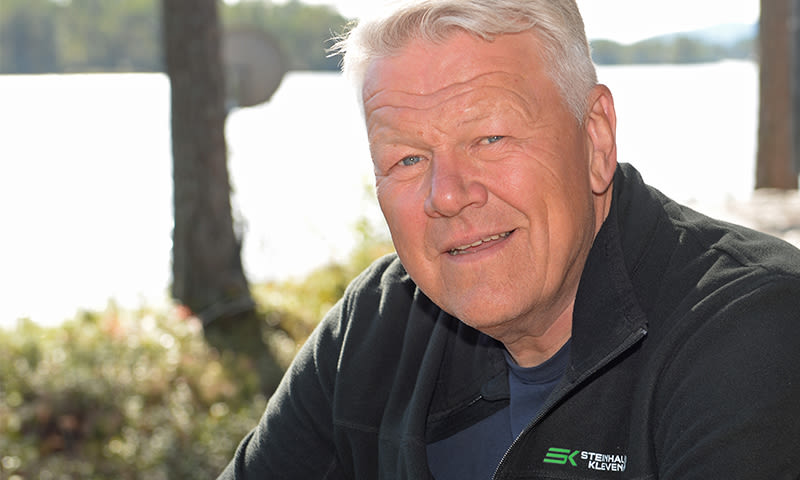 Mann i svart fleecejakke med grønn logo ved vannkant, trær og sollys i bakgrunnen.