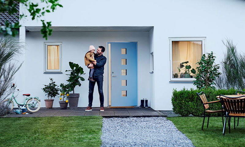 Far holder barn utenfor hvitt hus med blå dør, potteplanter, sykkel og hagemøbler