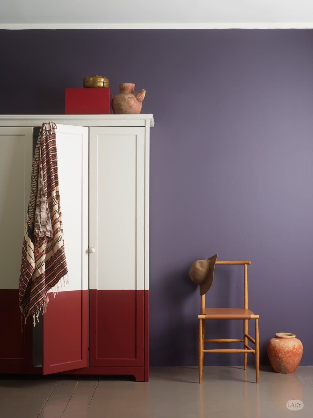 Hvit-rødt skap mot lilla vegg, stripet tekstil, trestol med hatt og terrakottapotter.