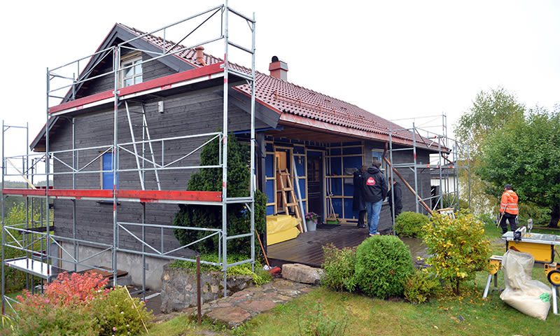 Grått trehus med rødt tak og stillas, håndverkere jobber i grønn hage.