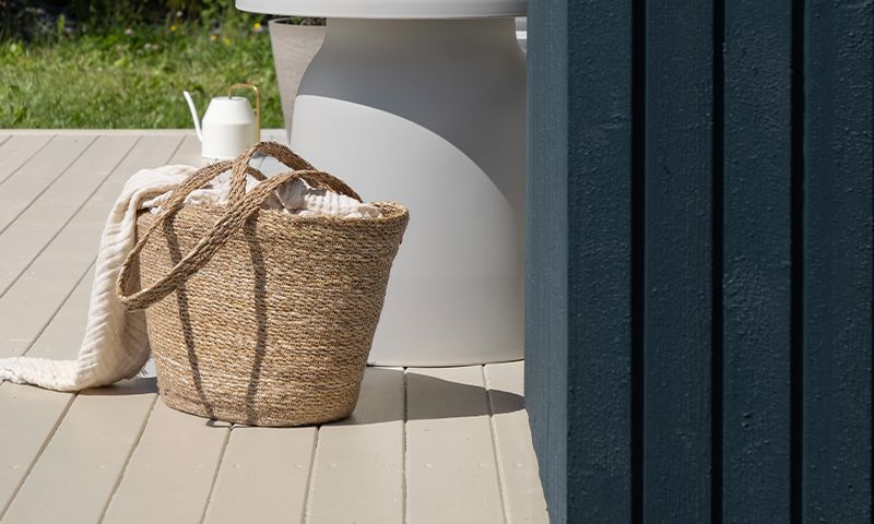 Beige flettet kurv med hvitt pledd på lyst terrassegulv ved hvitt bord og blå vegg.