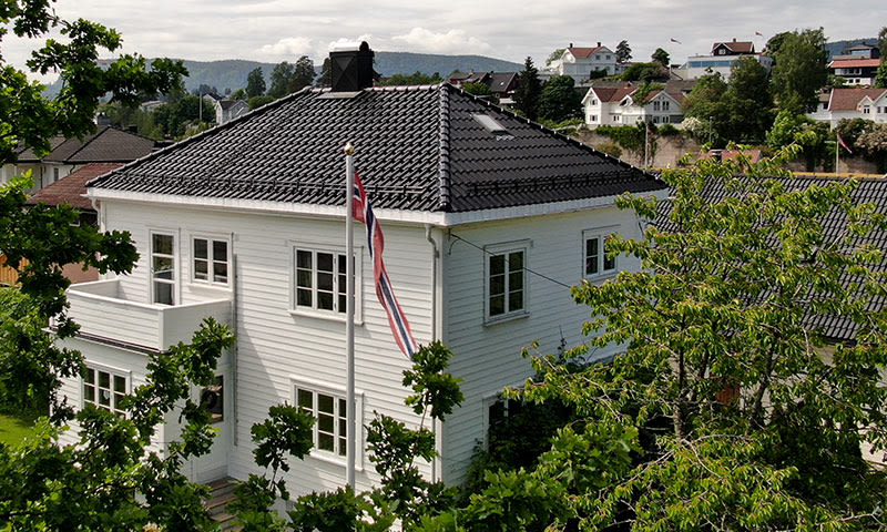 Hvit toetasjes villa med sort tak og norsk flagg, omgitt av grønne trær.
