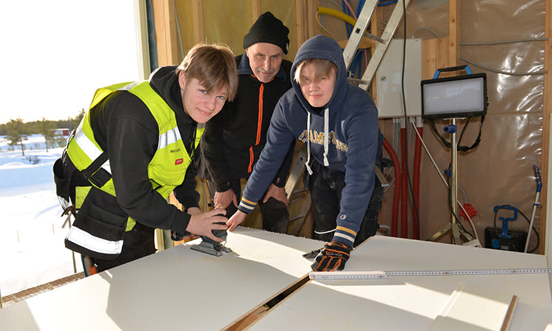 Håndverkere pusser gipsplate i uferdig bygg, én med gul refleksvest, synlige trebjelker