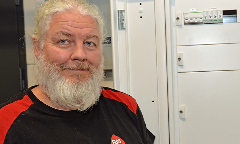 Mann med langt hvitt skjegg i svart-rød BM-t-skjorte foran hvite sikringsskap.