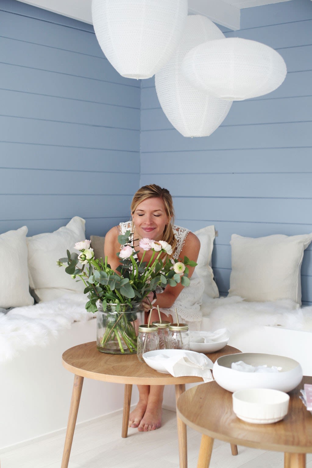 Hvitkledd kvinne ordner rosa og hvite ranunkler i glassvase på trebord mot lyseblå panelvegg.